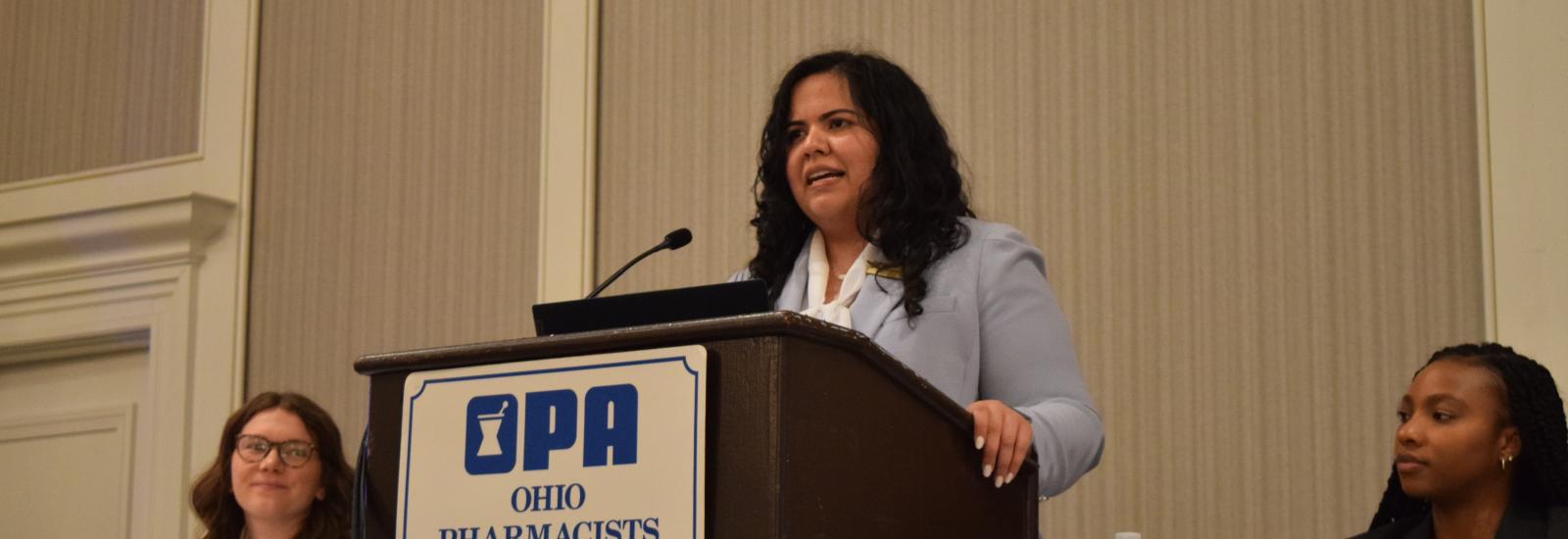 myriam speaking at a podium on OPA