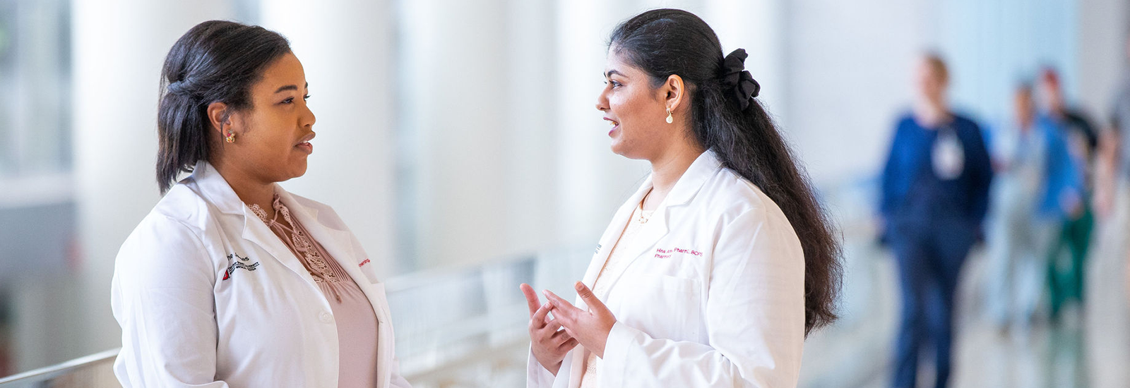 Two women in white coats at the hospital, conversing with each other.