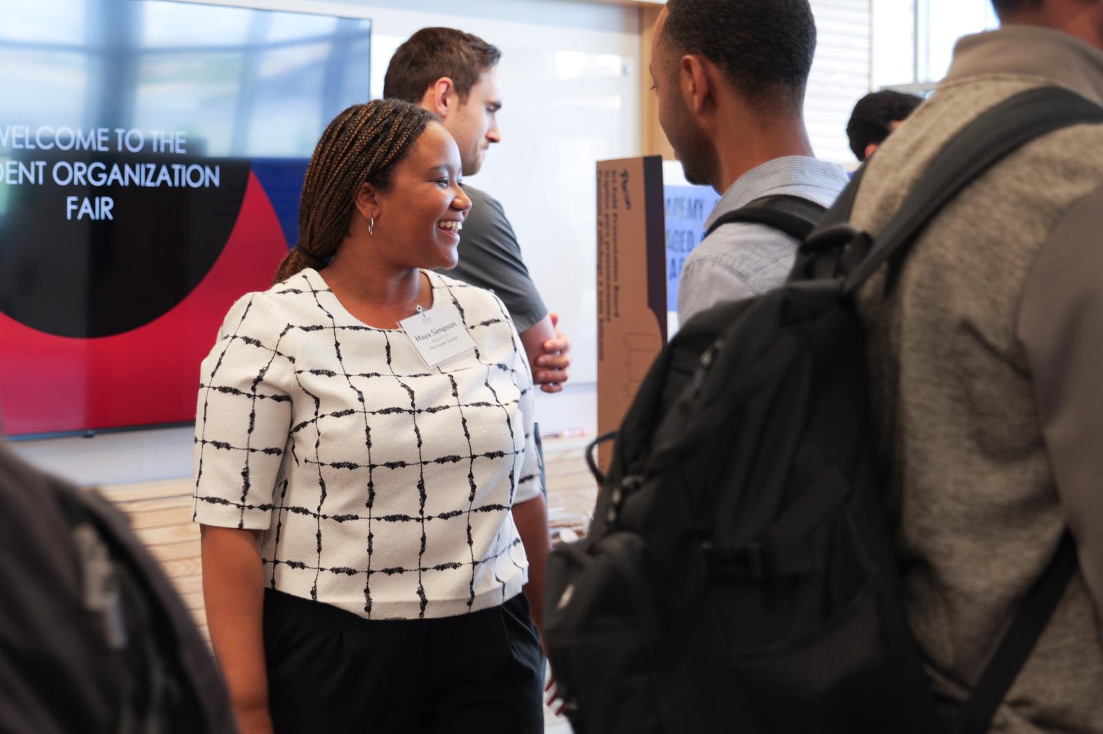 A student smiling at someone. There is a digital screen behind her that says in all caps "Welcome to the student organization fair" 