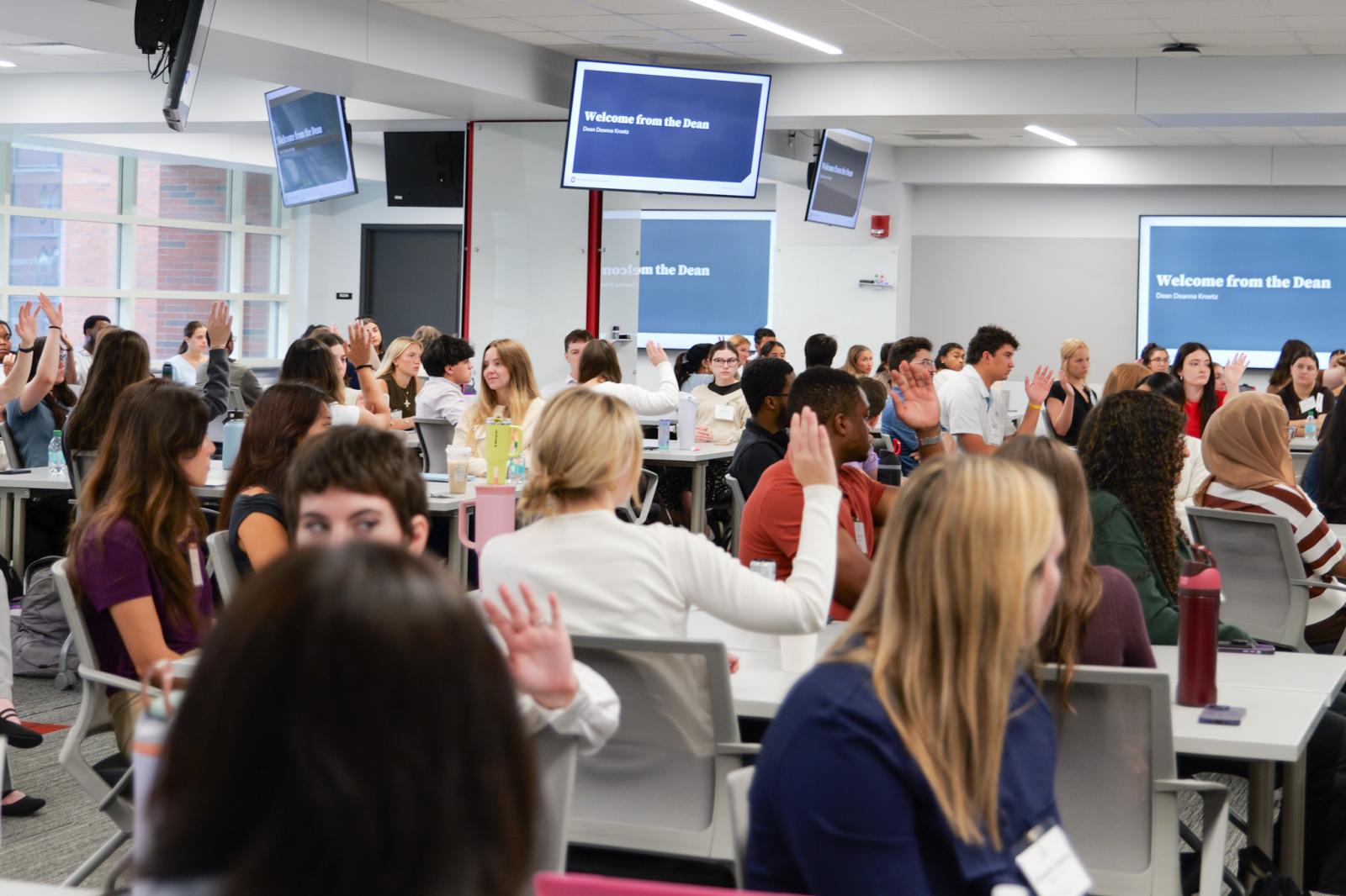 A large classroom of students holding up there hands to ask a question 