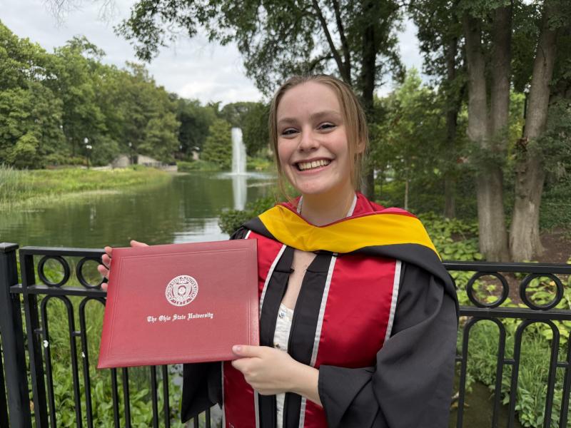 a person in regalia holding a diploma outside