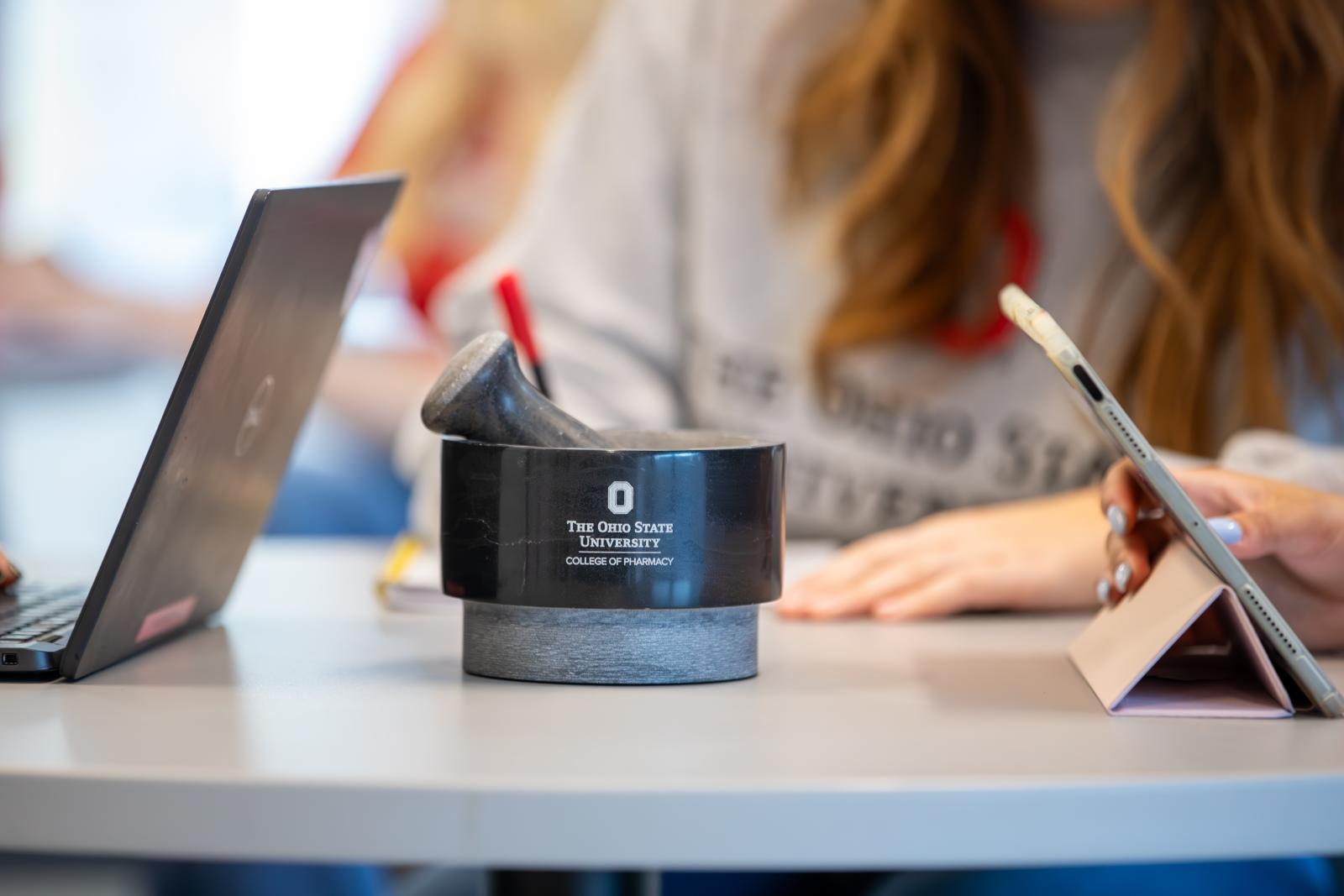 a mortar and pestle with college logo on table with students at laptops